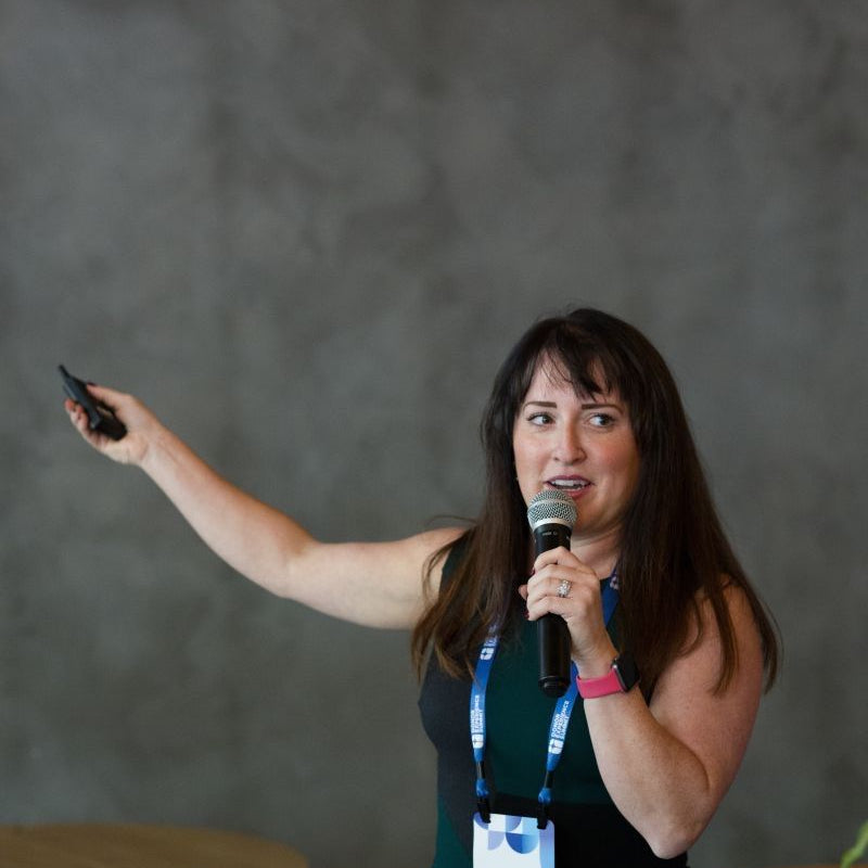 Therese, a woman with dark hair stands on a stage holding a microphone and pointing with a remote, wearing a dark green sleeveless dress and a conference badge.